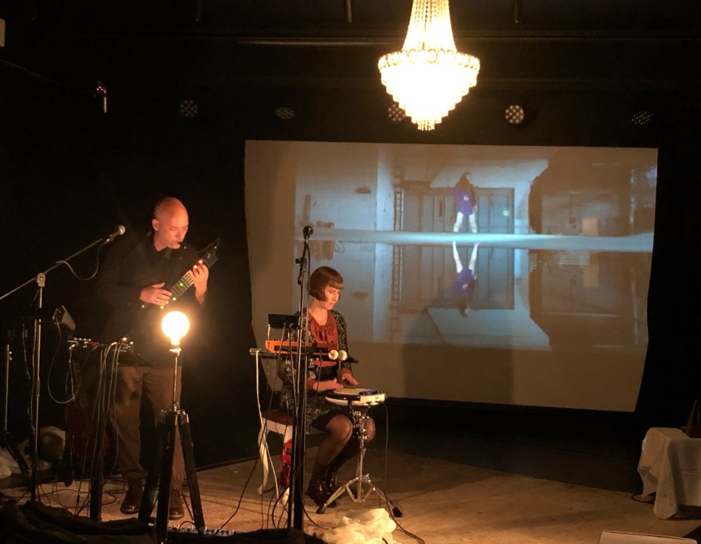 Man and woman playing instruments. Video on large screen in the background.